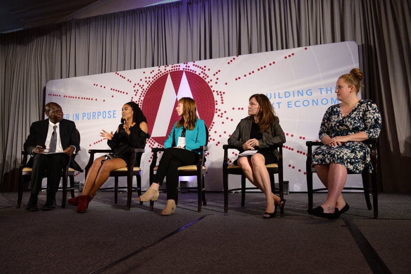 Panelists engaged in discussion seated in front of a custom fabric tension banner displaying Lead in Purpose and Building the Impact Economy