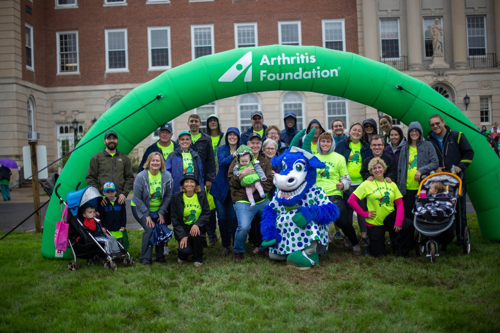 Group of participants and a mascot under a green Arthritis Foundation inflatable arch at a charity walk event.