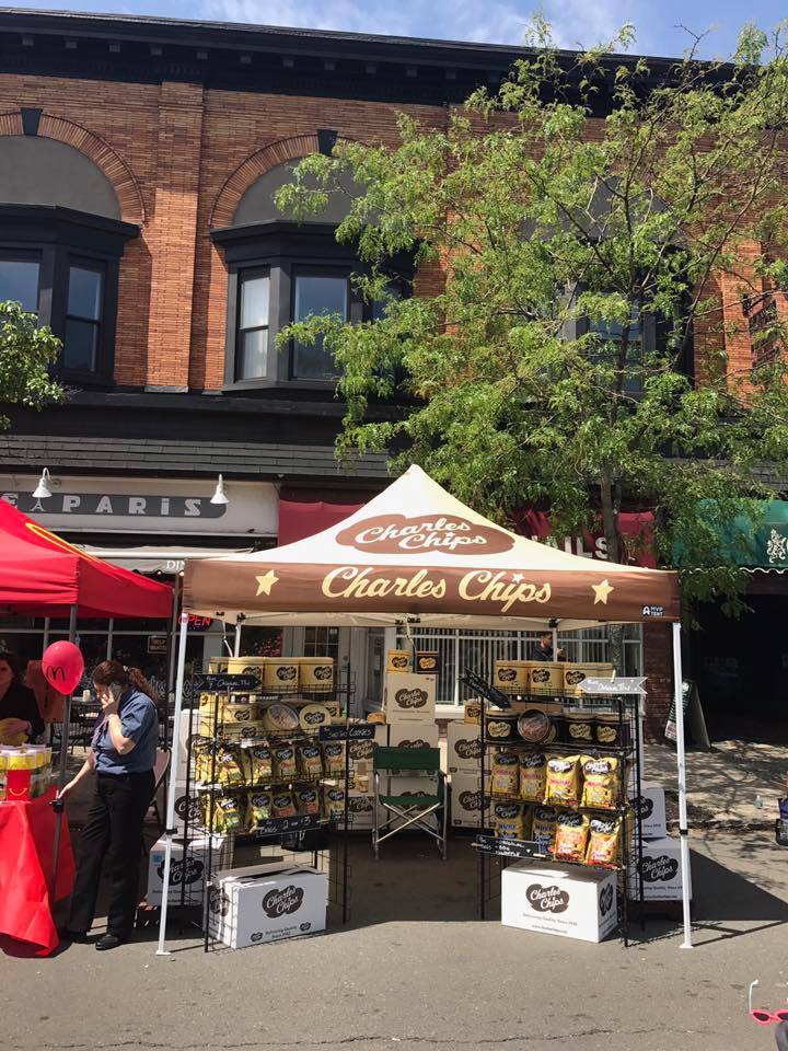 A Charles Chips farmers market 10x10 pop up tent displays an array of chip tins on a sunny day in a historic district 