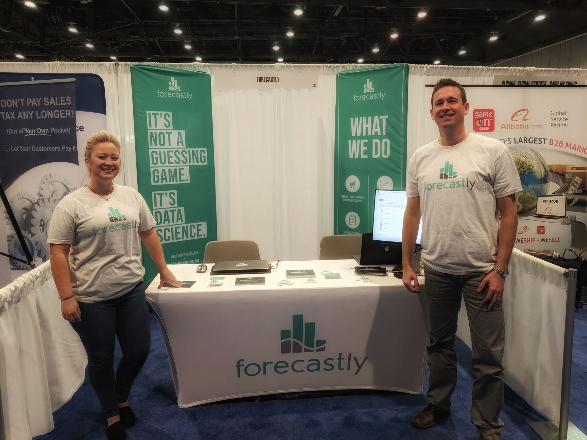 Trade show booth with Forecastly branded table cover, featuring company logo and tagline, with two smiling representatives