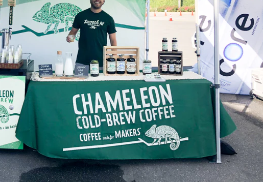 Green trade show table cover with Chameleon Cold-Brew Coffee logo, showcasing products and engaging with attendees
