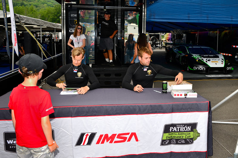 a car racing team autograph session featuring a custom outdoor tablecloth with the IMSA logo and racing car in the background