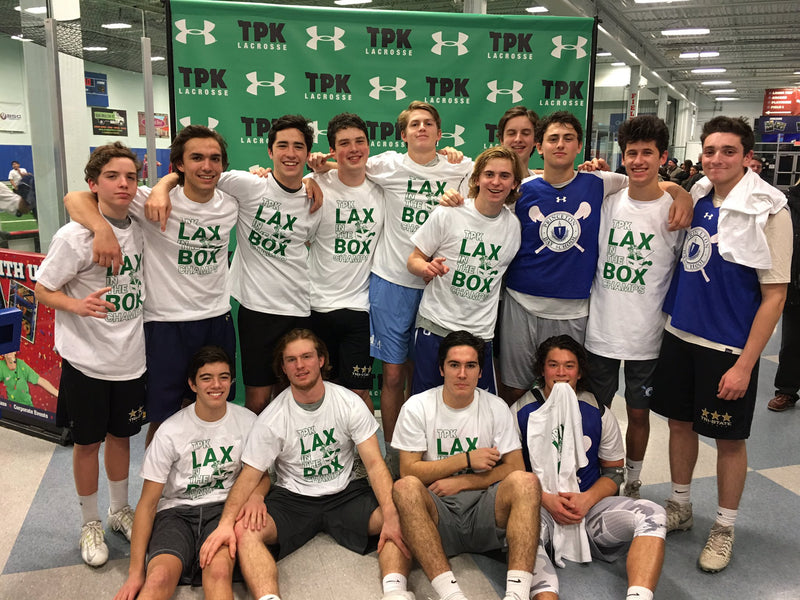 Lacrosse team posing with trophy in front of a branded photo backdrop at indoor sports facility.