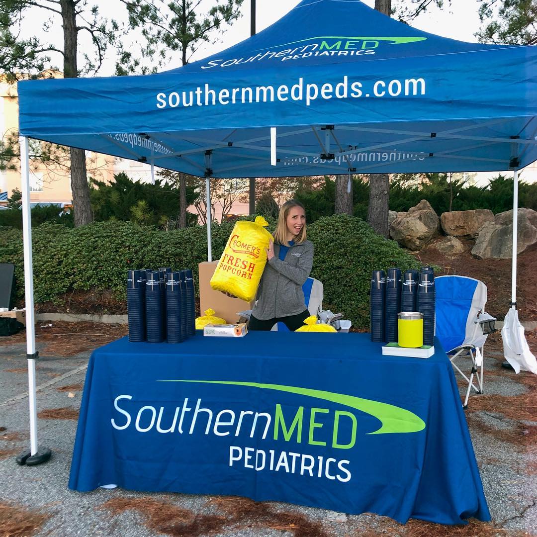 A smiling attendant stands under a blue 10x10 trade show tent with logo, with a table and promotional materials ready
