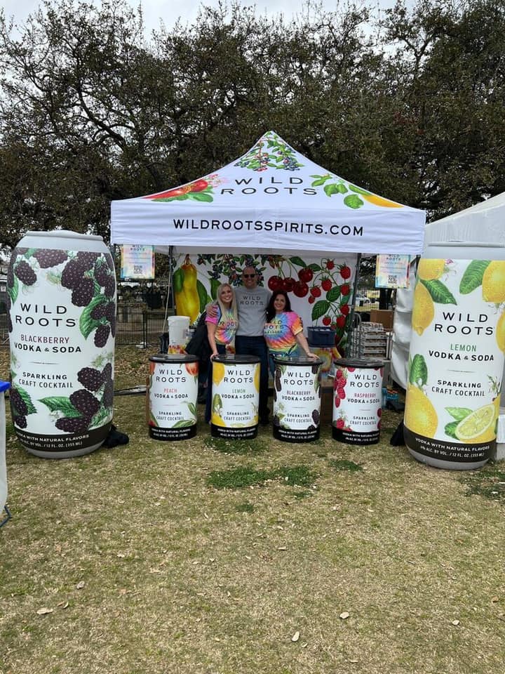 Enthusiastic vendors pose at a custom farmers market pop up tent adorned with vibrant fruit designs for Wild Roots Spirits