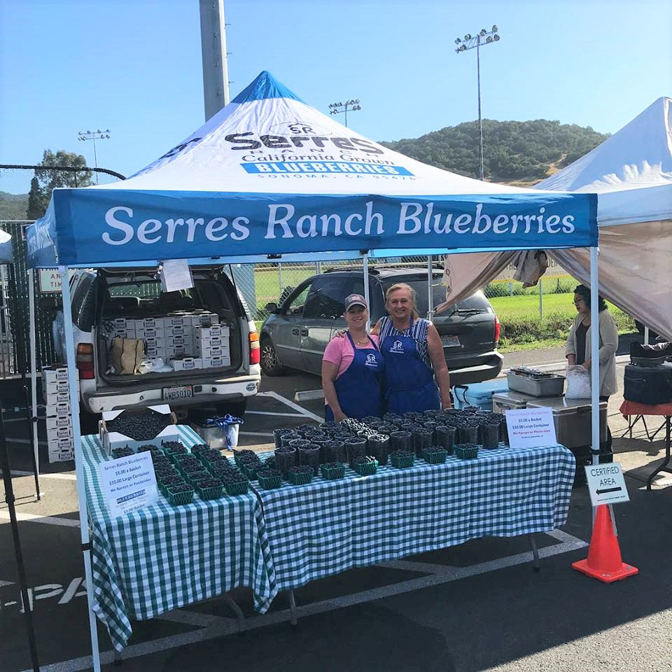 Smiling vendors stand by a farmers market tent labeled Serres Ranch Blueberries under a clear blue sky