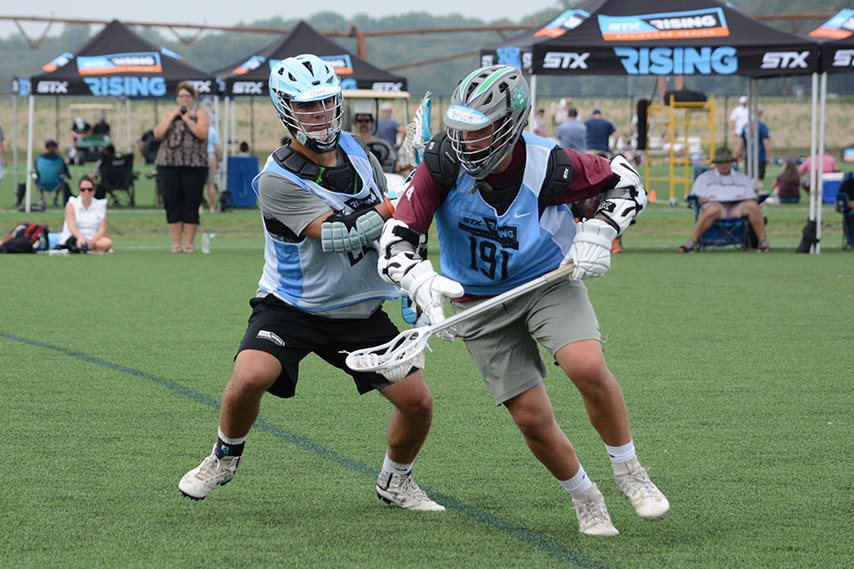 two lacrosse players wrestling for a superior position with a line of lacrosse branded canopy tents in the background