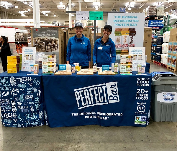 Perfect Bar branding table set up in a store, with two smiling representatives in matching outfits presenting the product range and key benefits listed on the tablecloth