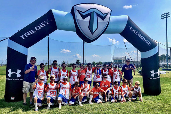 Youth lacrosse team posing under a black Trilogy Lacrosse custom shaped inflatable arch with Under Armour logos