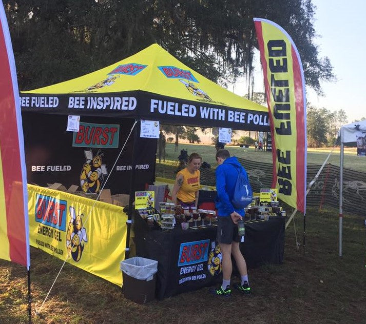 A vibrant farmers market vendor tent with BEE FUELED branding offers energy gels to customers in a grassy area