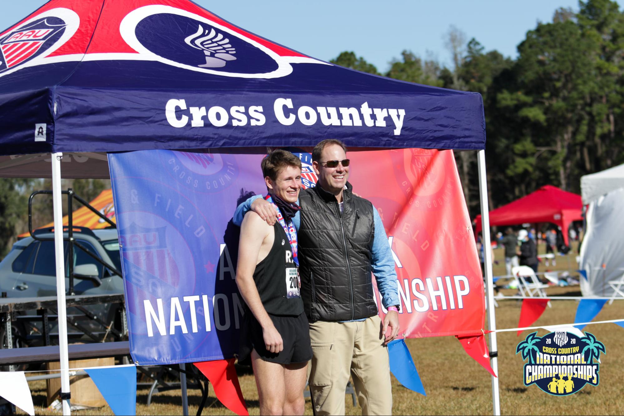 Cross Country event tent with a custom printed canopy and backdrop banner at an outdoor national championship. A runner wearing a medal poses with an event official under the tent, with other tents and event attendees visible in the background.