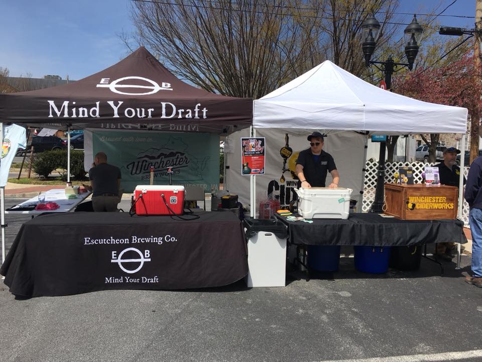 Outdoor booth setup featuring a brown Mind Your Draft canopy for Escutcheon Brewing Co. and a white canopy for Winchester Ciderworks at a local event