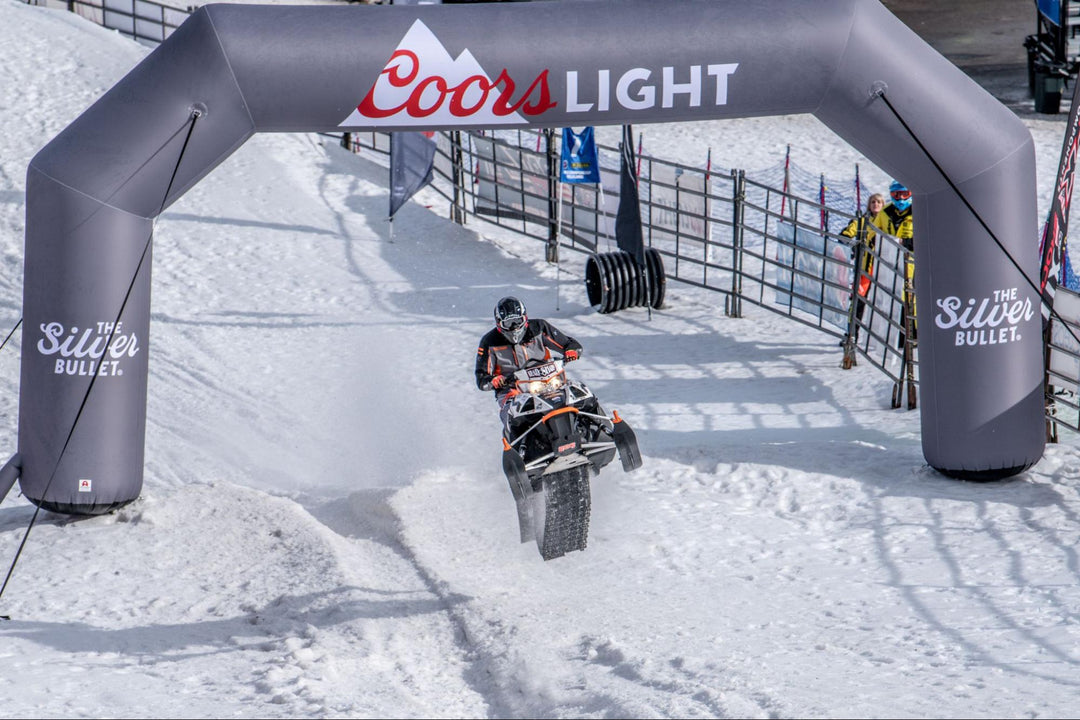 A snowmobile races under a branded Coors Light inflatable arch at a snowy outdoor event.
