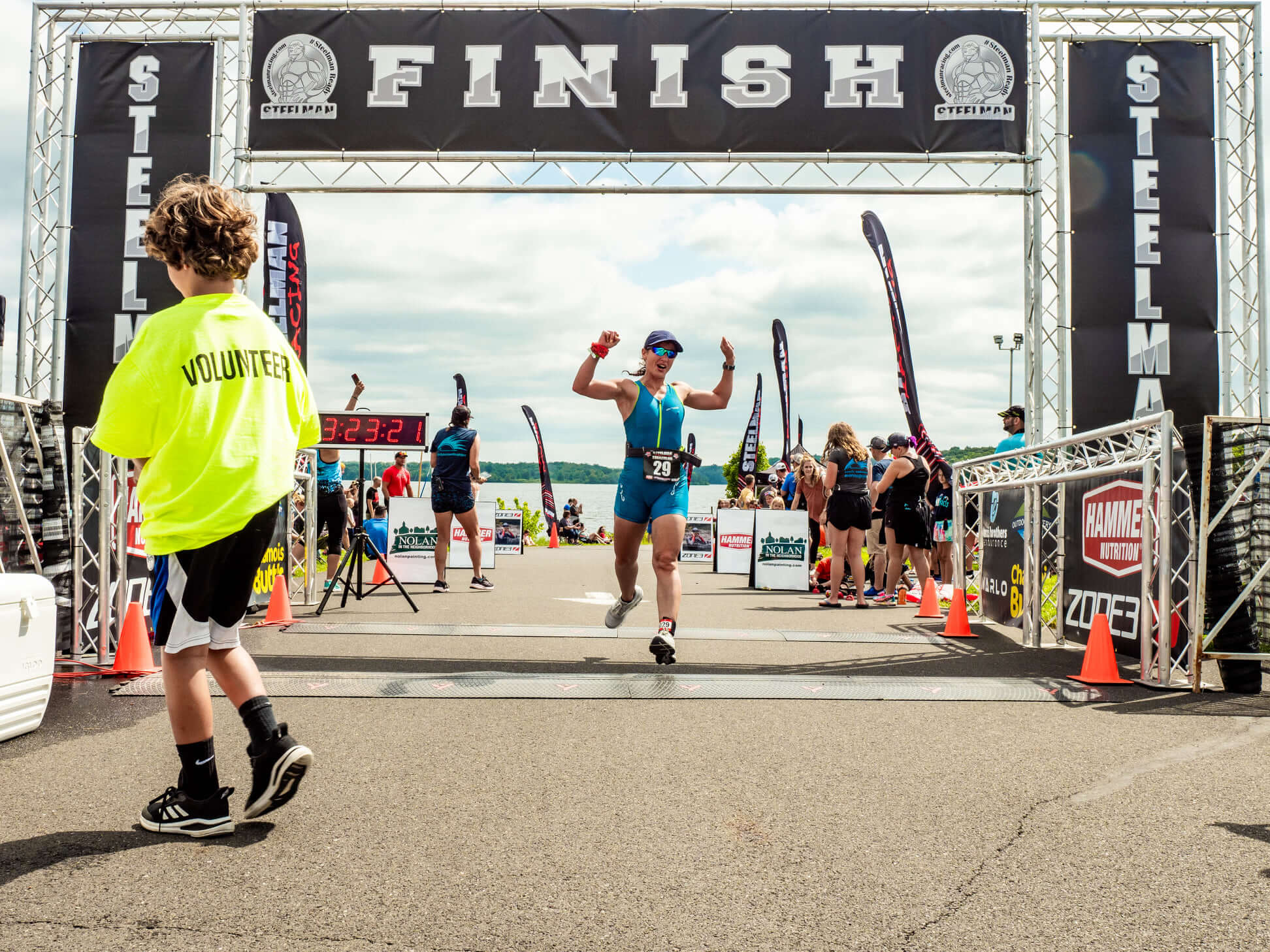 Athlete crossing the finish line under a large "FINISH" banner at a Steelman race event, with a volunteer in a yellow shirt nearby and spectators in the background.