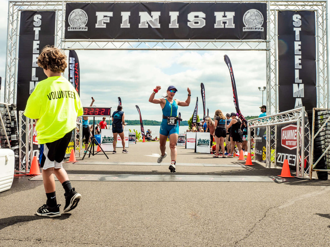 Athlete crossing the finish line under a large "FINISH" banner at a Steelman race event, with a volunteer in a yellow shirt nearby and spectators in the background.