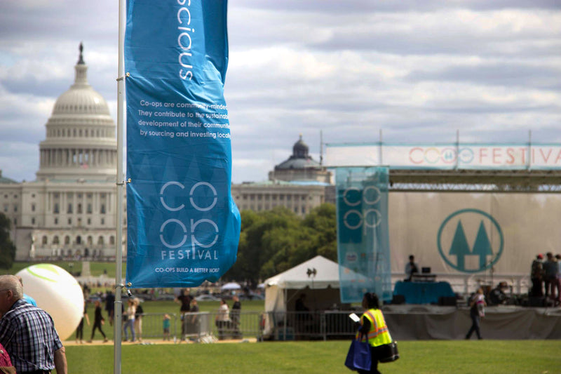 A blue CO-OP FESTIVAL customized feather flag in focus with the Capitol building blurred in the background at an outdoor event