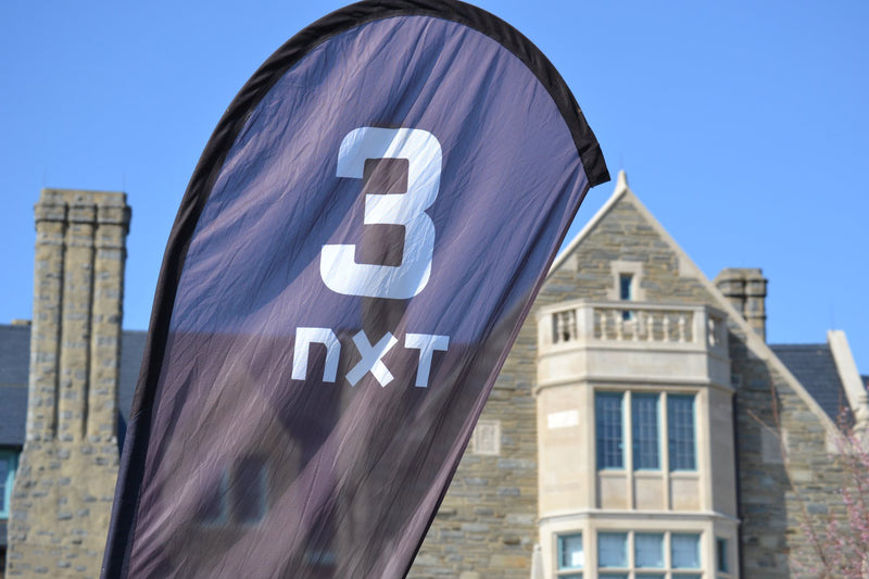 Custom printed teardrop flag with 3 NXT branding in front of a stone building under blue sky