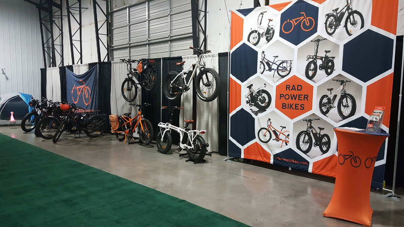 Exhibit of Rad Power electric bikes with a custom backdrop with stand, promotional materials on display, indoor event.