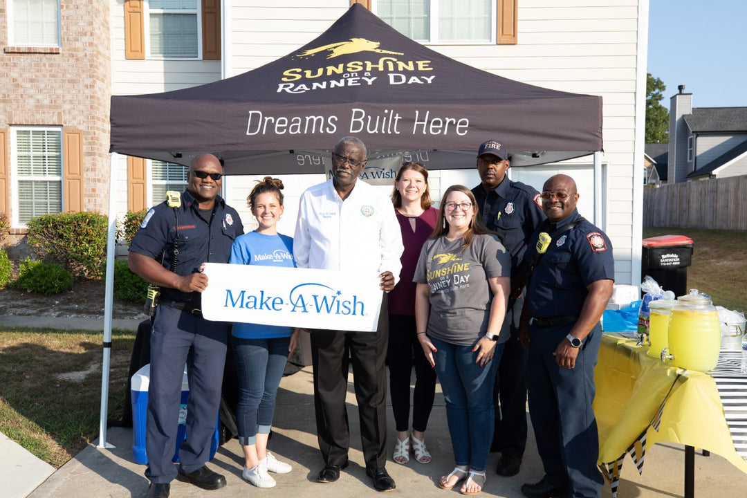 Community members and firefighters gathered under a Sunshine on a Ranney Day event canopy holding a Make-A-Wish sign.