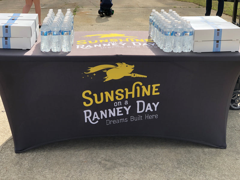 A promotional table covered with a Sunshine on a Ranney Day - Dreams Built Here branded tablecloth, displaying water bottles and boxes.