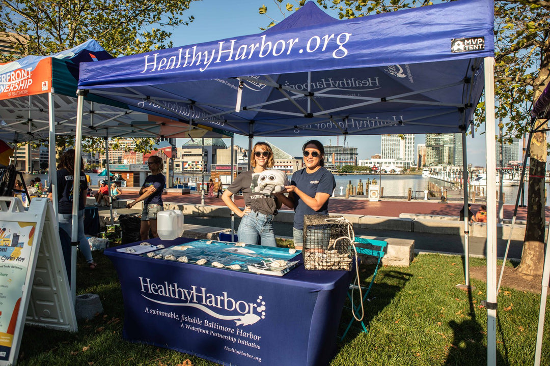 Two volunteers at a HealthyHarbor.org nonprofit booth under a custom canopy at a waterfront event in Baltimore.