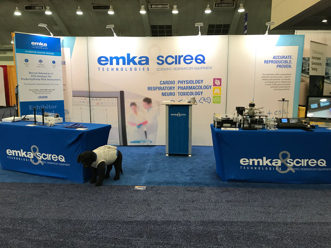 Trade show booth for Emka Technologies with informative backdrop banners displaying scientific respiratory equipment and a plush dog model