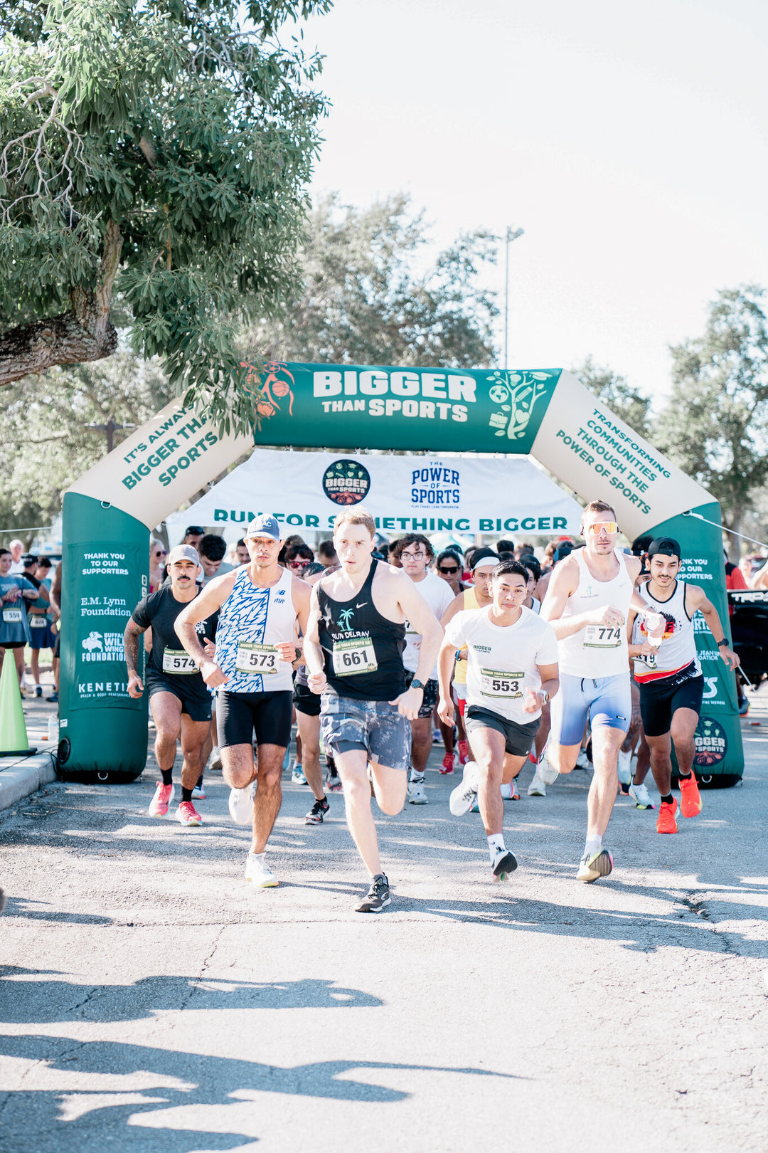 Group of male runners starting a race under a "Bigger Than Sports" inflatable archway during a daytime event