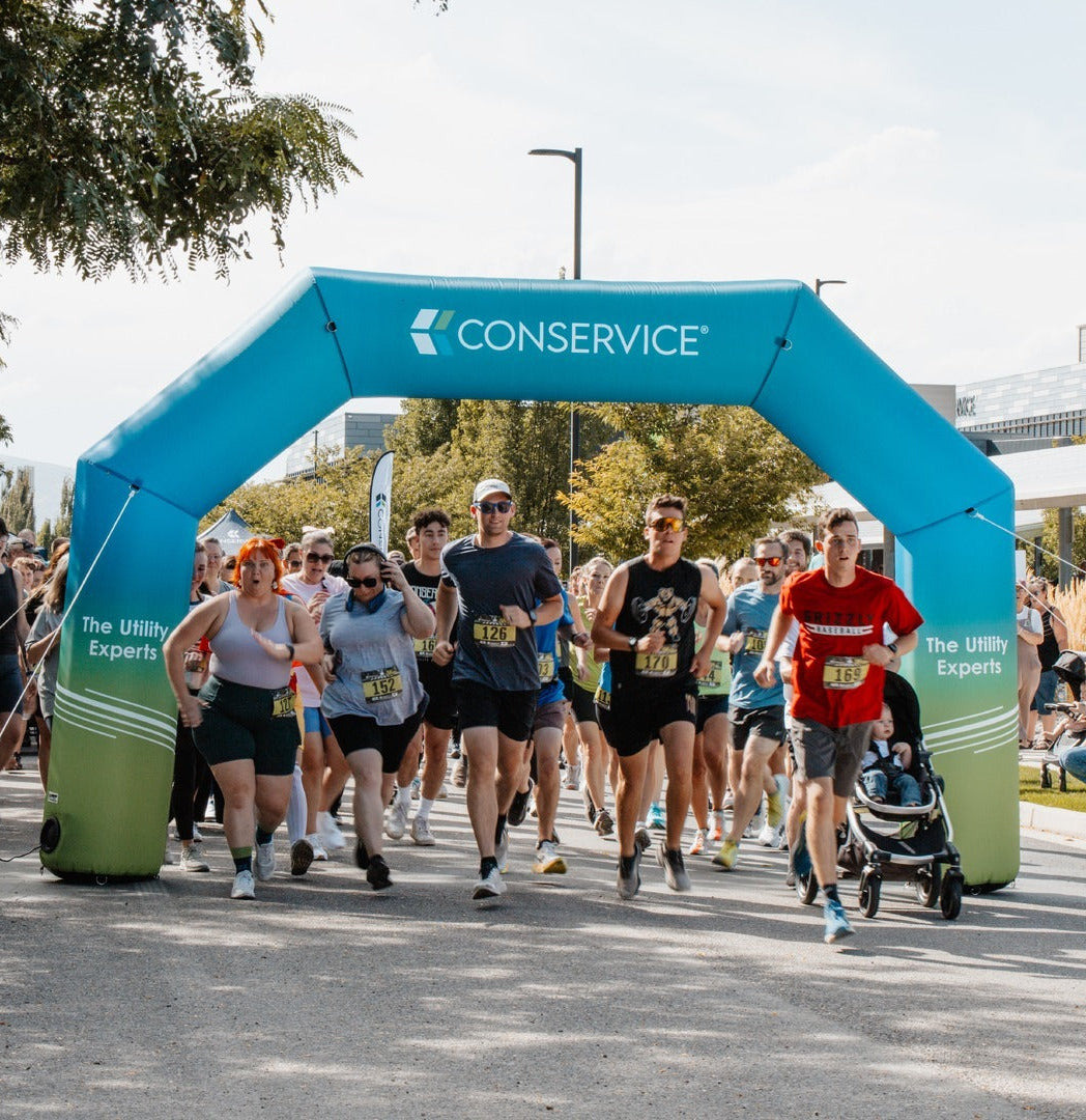 Group of runners starting a race under a branded blue and green Conservice inflatable arch at an outdoor community event