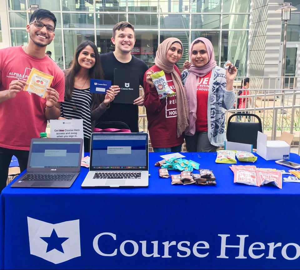 Group of people standing behind a table with laptops and promotional materials, branded 'Course Hero'.