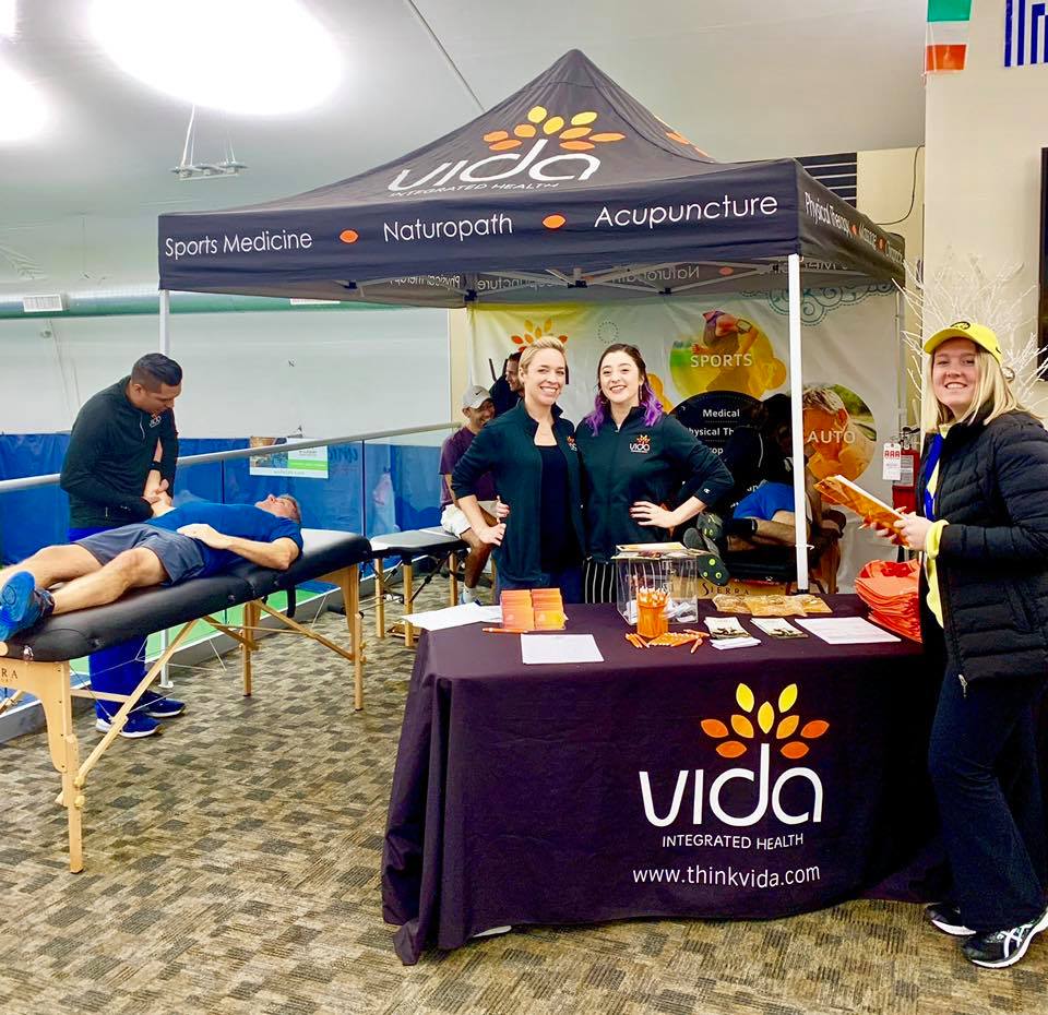 Custom branded table cover for Vida Integrated Health at wellness event booth with staff and treatment area