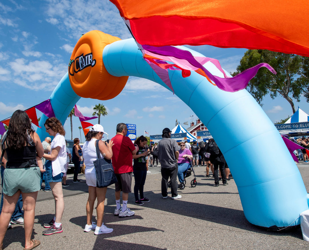 Wavy light blue inflatable arch with orange circular logo and colorful flags at OC Fair entrance