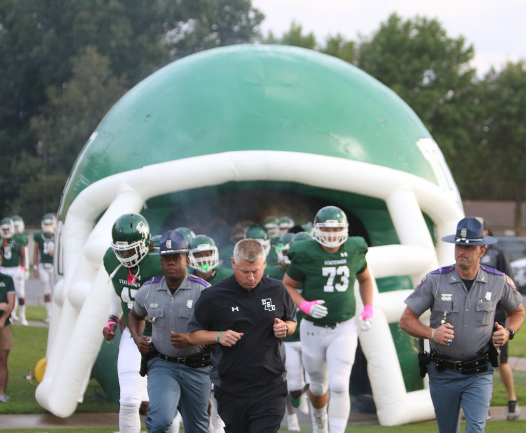 football players running out of inflatable green helmet tunnel with police escort