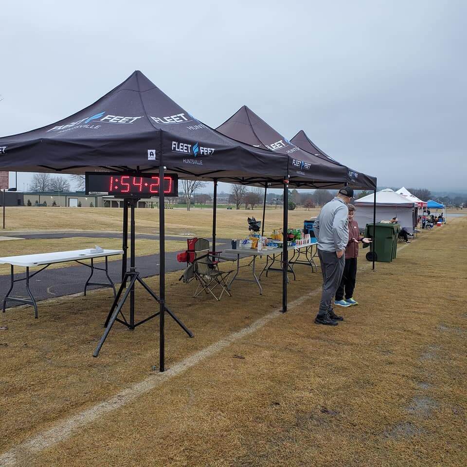 Fleet Feet Huntsville branded canopy tents at outdoor race event with event timer, tables, and refreshments