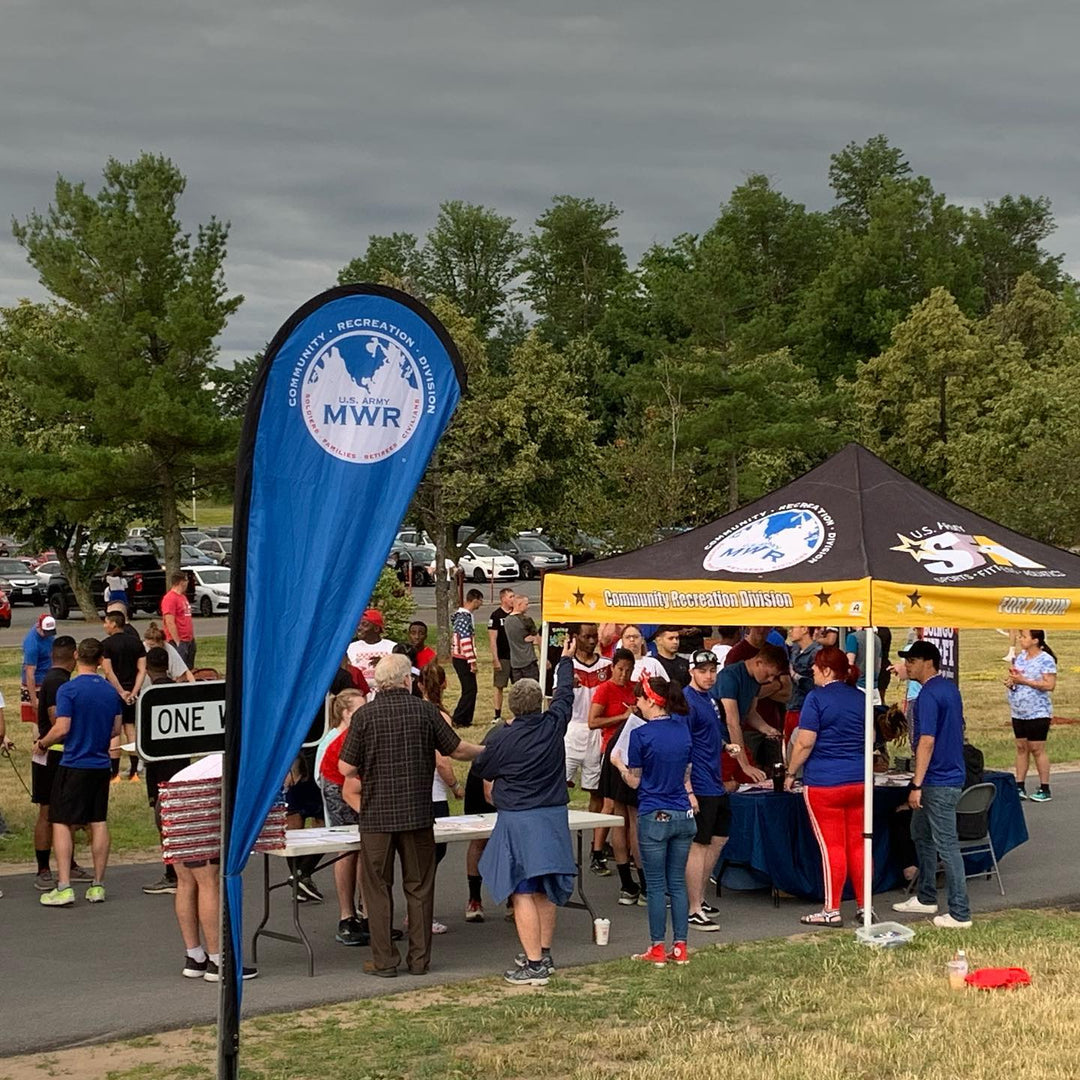 Blue U.S. Army MWR feather flag at Fort Drum community recreation event with crowd near branded tent.
