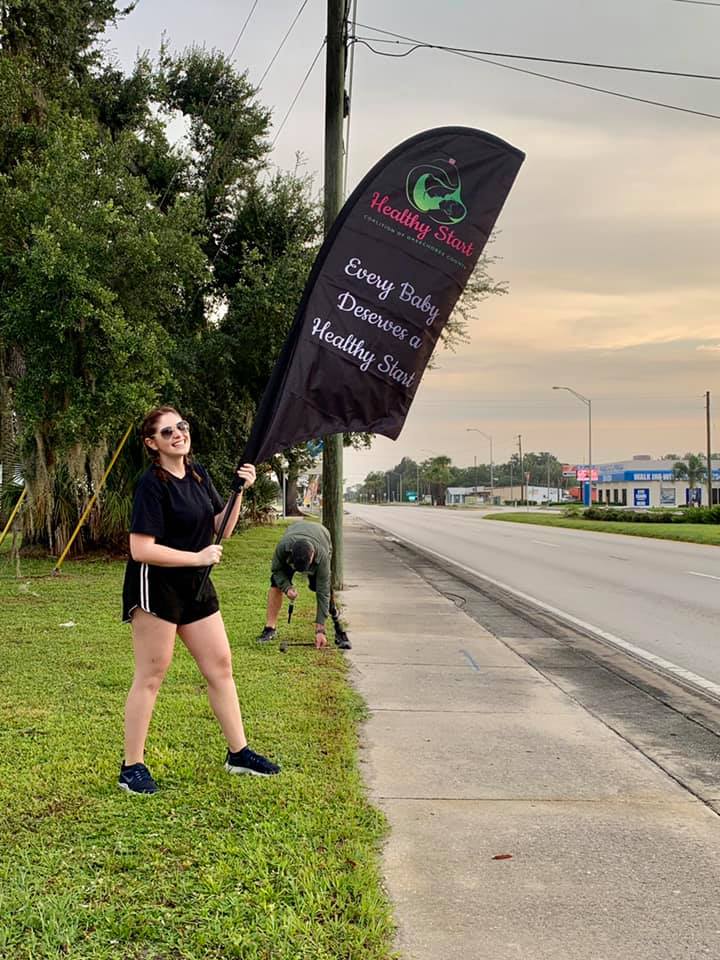 Woman holding a black feather flag for Healthy Start with the slogan "Every Baby Deserves a Healthy Start" on a grassy roadside.