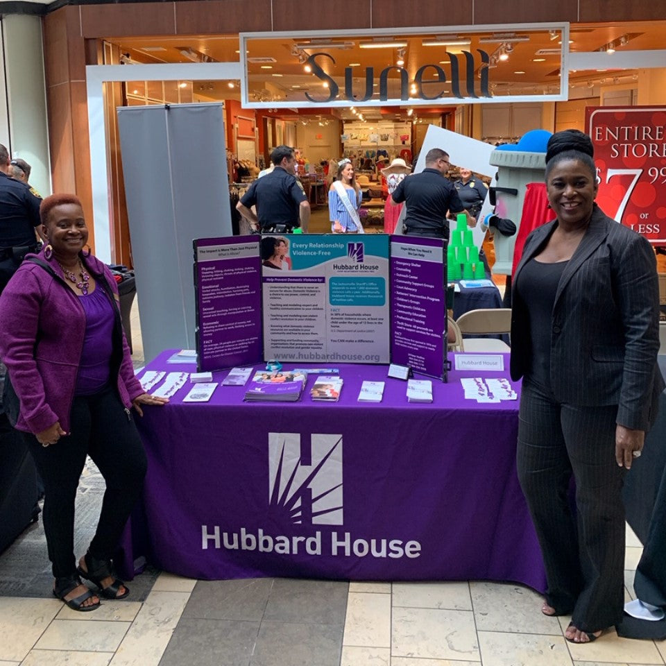 Two women standing next to a Hubbard House table at an event with a 'Surell' booth in the background.