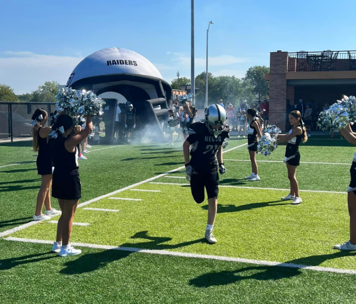 High school football player running onto the field through a 'RAIDERS' inflatable helmet tunnel with cheerleaders on the sides