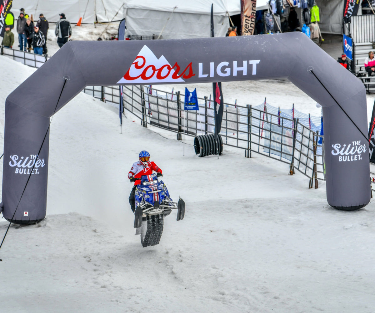 Snowmobile rider airborne during a winter race under a custom inflatable arch with Coors Light and The Silver Bullet branding