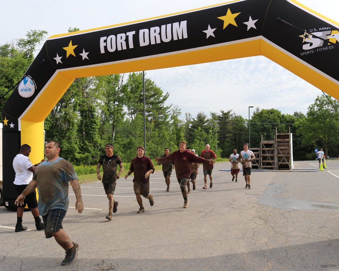 Participants completing a muddy obstacle course under a custom inflatable arch branded for Fort Drum and U.S. Army Sports Fitness and Aquatics, built by MVP Visuals