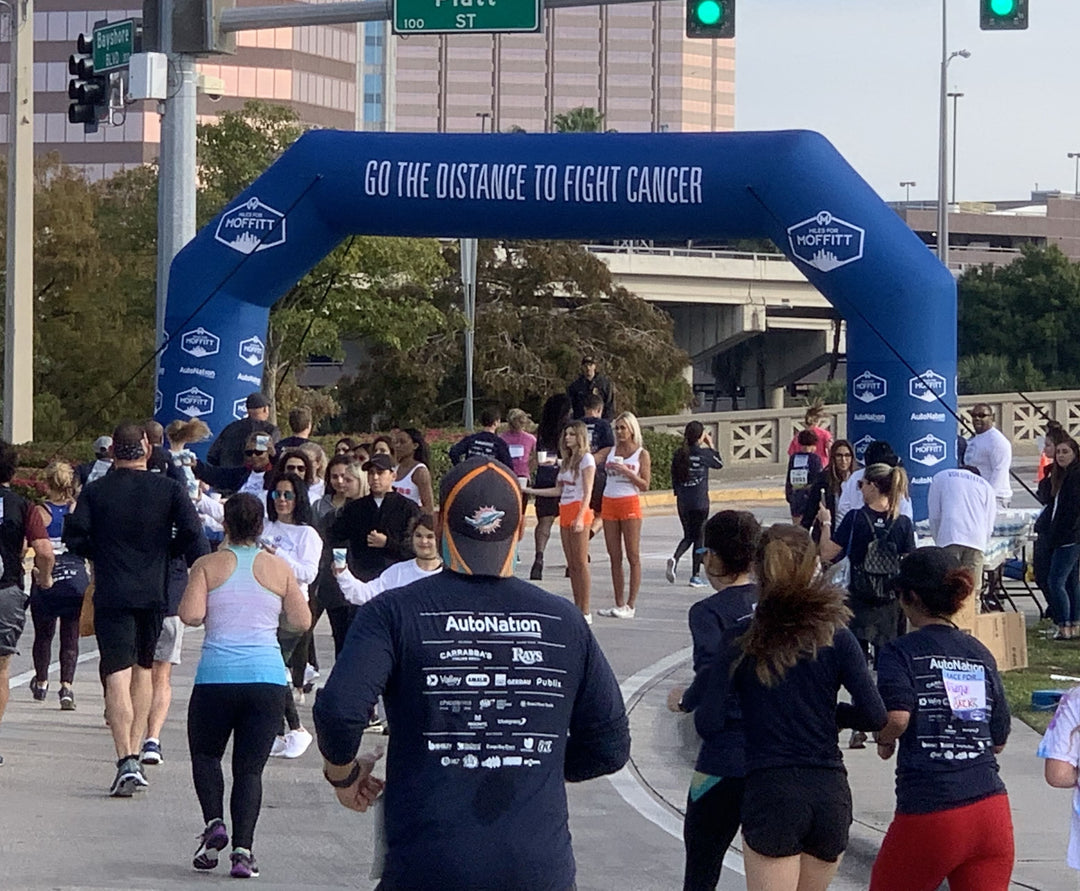 Runners passing through a custom inflatable arch with Moffitt Cancer Center branding and the message Go the Distance to Fight Cancer at a charity race