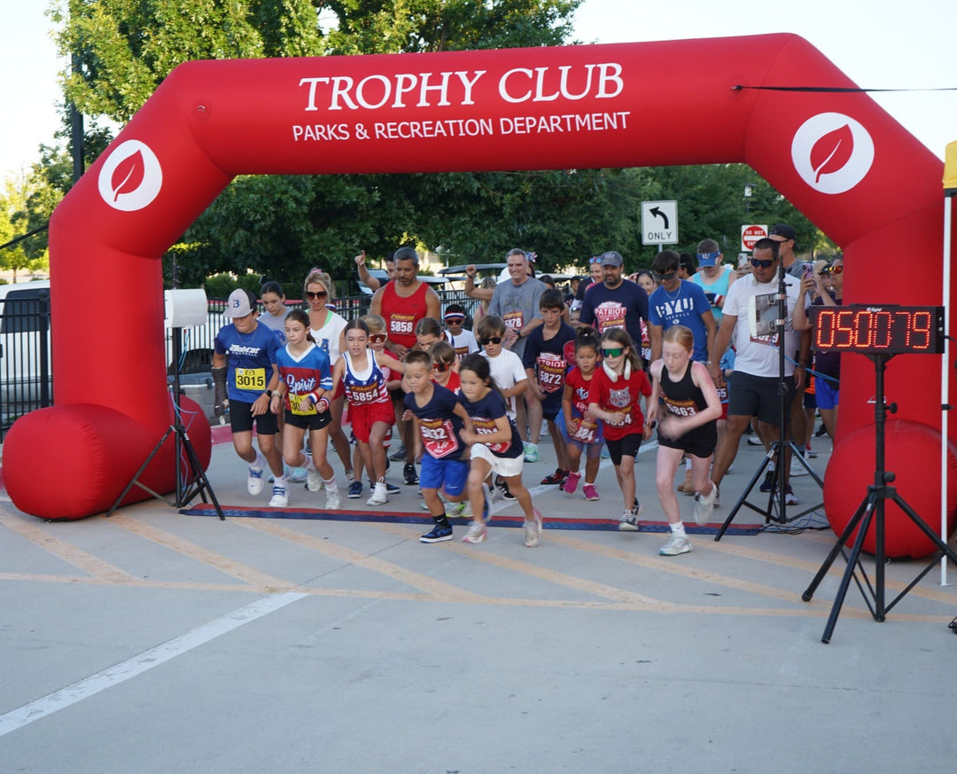 Children sprinting from the start line under a bright red inflatable arch for Trophy Club Parks and Recreation Department at a community fun run