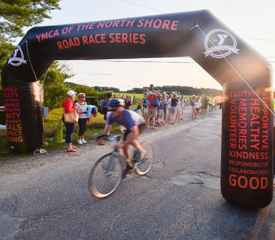 Cyclist racing under a custom black inflatable arch for YMCA of the North Shore Road Race Series as spectators cheer along a scenic route at sunset