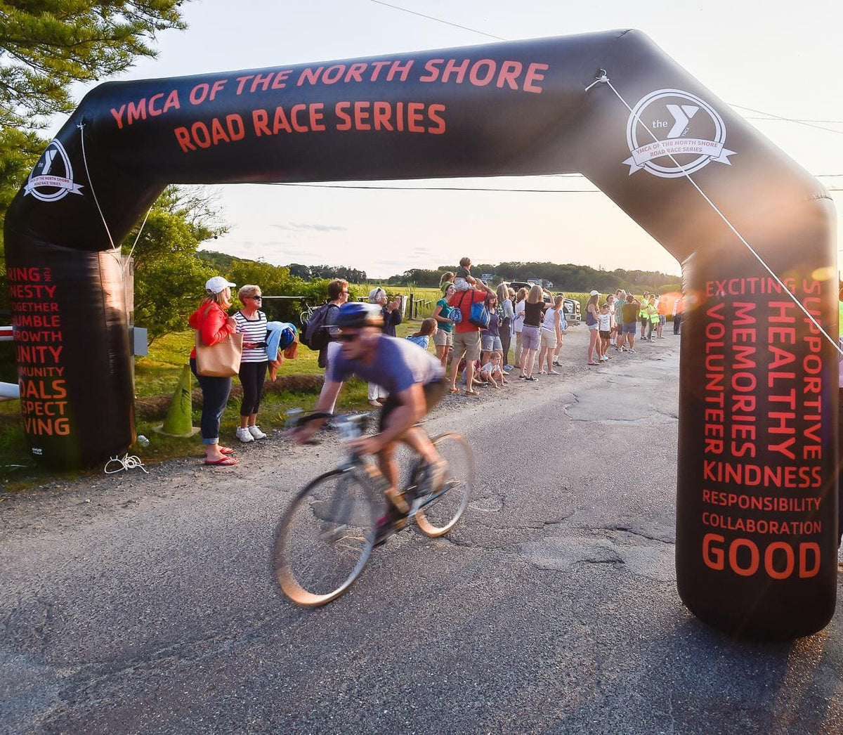 Cyclist racing under a custom black inflatable arch for YMCA of the North Shore Road Race Series as spectators cheer along a scenic route at sunset