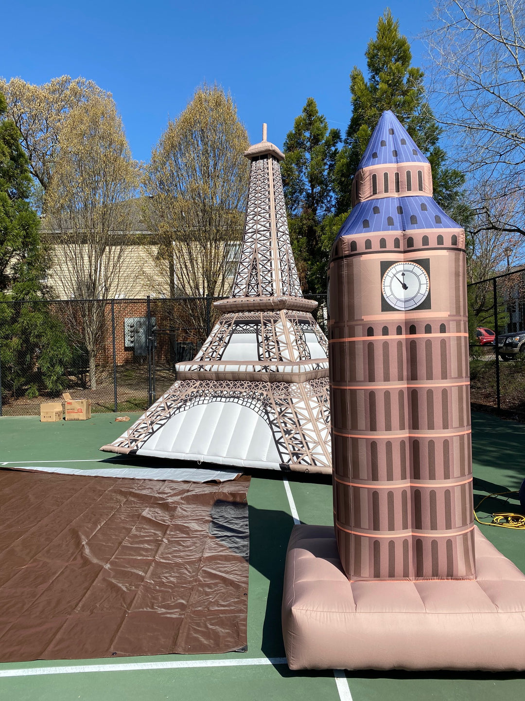 Inflatable replicas of the Eiffel Tower and Big Ben standing outdoors on a tennis court