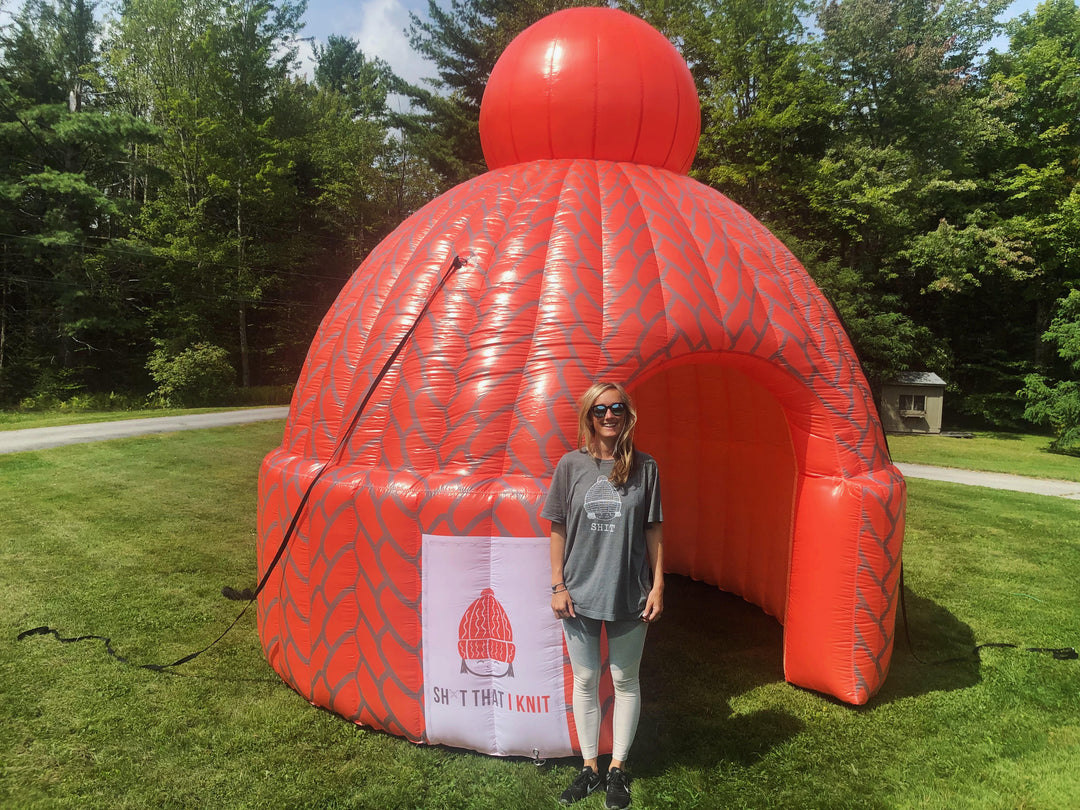 Inflatable red knit hat dome with "Sh*t That I Knit" branding on grassy outdoor field
