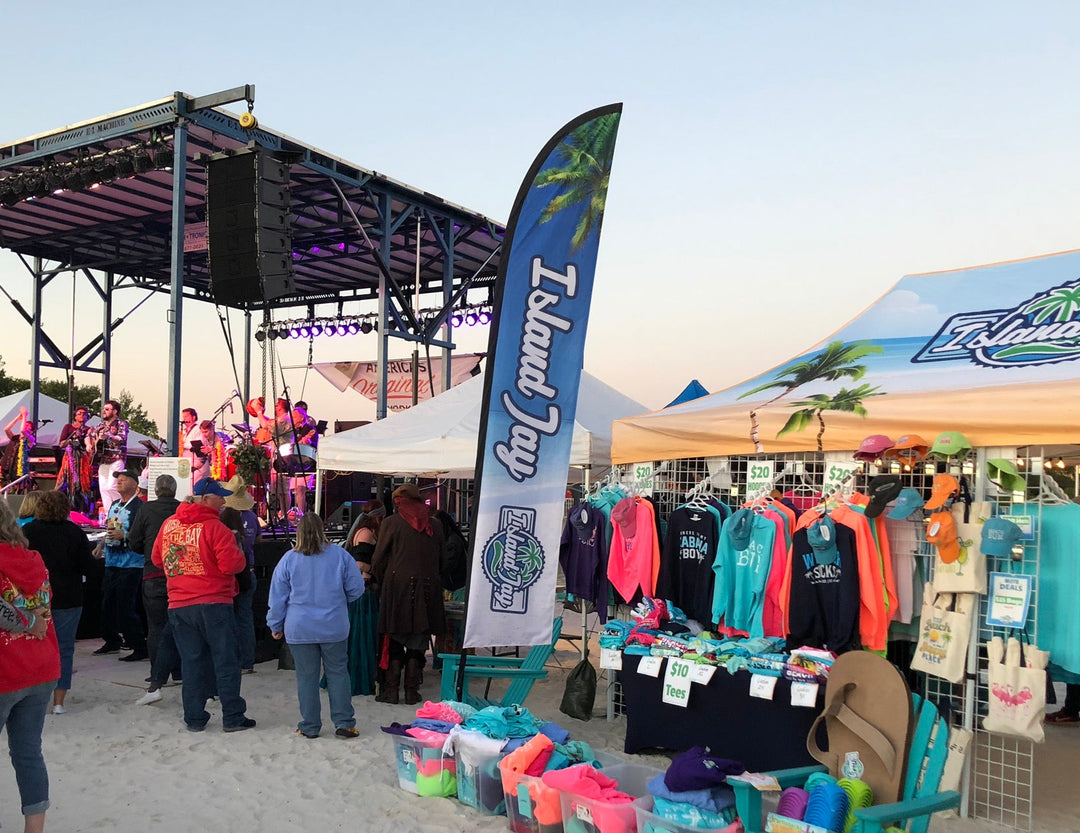Island Jay feather flag and beachwear merchandise booth next to a live band performance on the sand at an outdoor event.