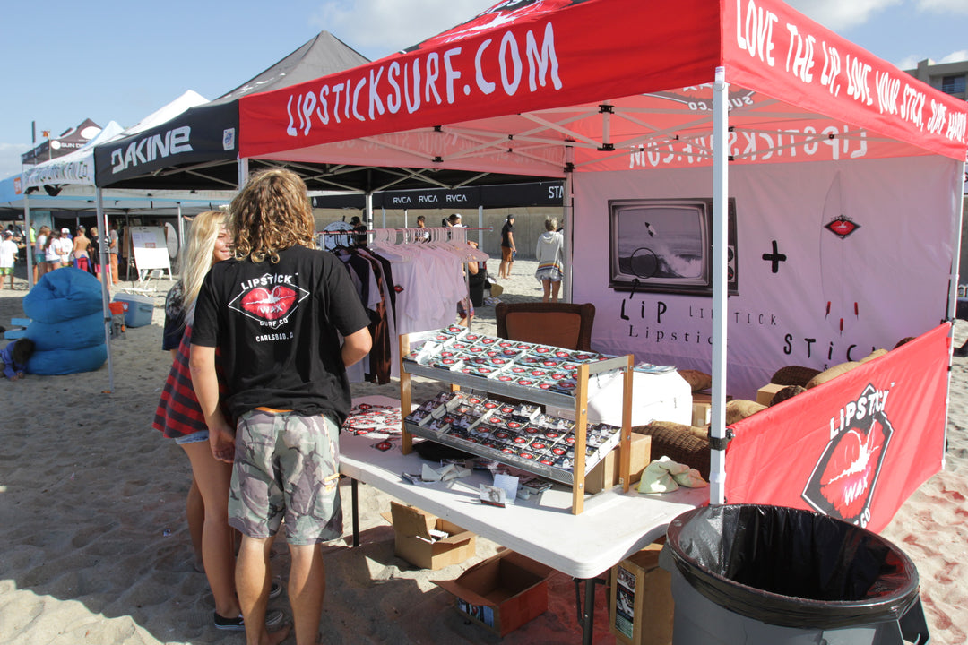 People at a beach event with a Lipstick Surf tent and merchandise display.