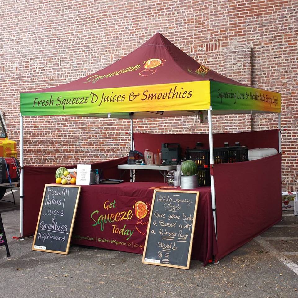 A maroon and yellow farmers market canopy offers fresh juices and smoothies against a brick wall backdrop