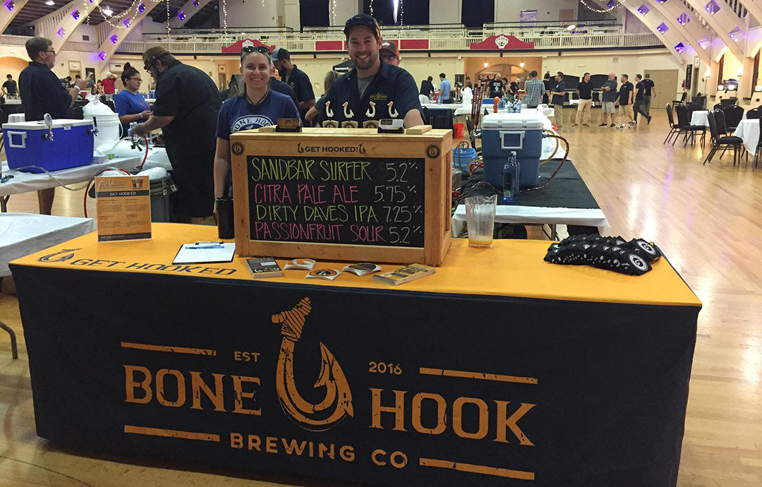 Bone Hook Brewing Co booth at an event, featuring a logo table cover, beer list, and smiling staff