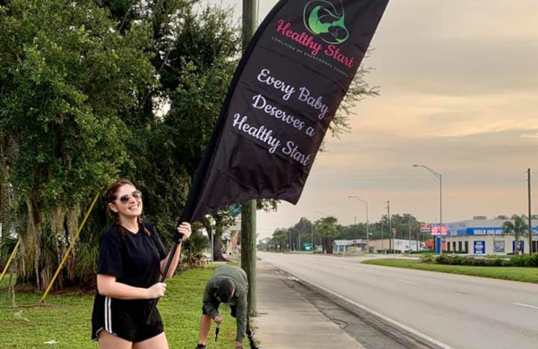 Woman proudly holding a custom printed feather flag promoting Healthy Start with the slogan Every Baby Deserves a Healthy Start on the roadside